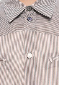 Pruhovaná košile hnědé a bílé barvy, se špičatým límcem, dvěma knoflíky a kapsou s detailem šikmého prošití. Měkká textura látky.