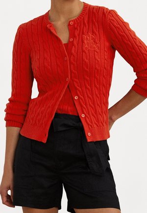 Vrouw draagt een rode kabelgebreide cardigan met knopen en bijpassend mouwloos topje, gecombineerd met zwarte hoge taille shorts met een stoffen riem.