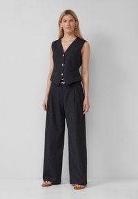 Mouwloze donker gestreepte vest met gouden knopen en twee zakken, gecombineerd met een breedvallige gestreepte broek. Platte sandalen maken de outfit af.