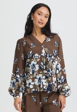 Femme portant un chemisier marron et un pantalon avec de grands motifs floraux blancs et bleu marine, debout sur un fond clair uni.