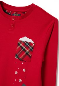 Camicia rossa a maniche lunghe con due bottoni frontali, taschino a quadri sul petto, design in schiuma bianca all'interno della tasca e fiocchi di neve bianchi sparsi in basso.
