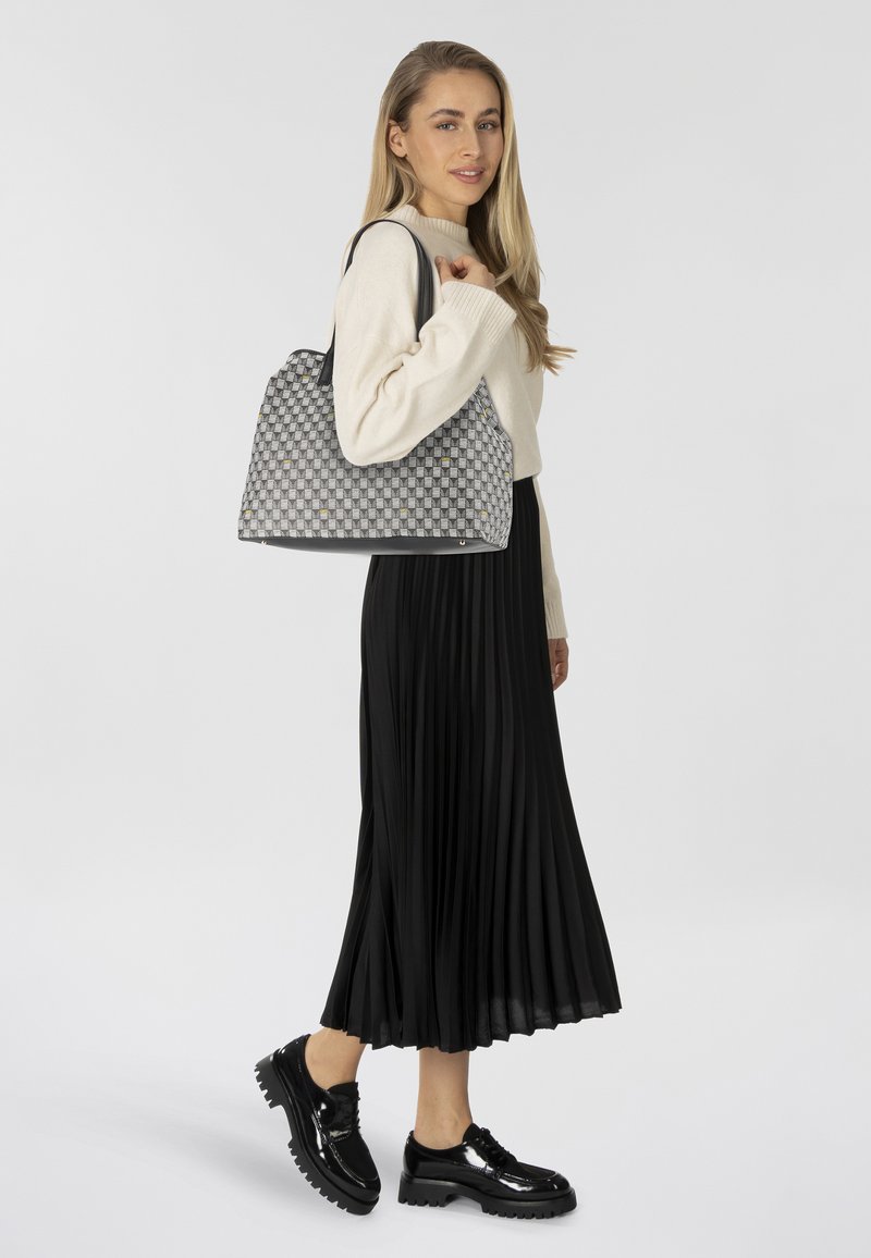 Grijze gevlochten handtas met een geruit patroon, zwarte leren schouderbanden en een matte afwerking. Model draagt een geplooide zwarte rok en lakleren schoenen.