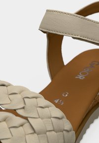 Φωτεινό μπεζ πλεκτό δερμάτινο σανδάλι με λουράκι στον αστράγαλο, διαθέτει λεία καφέ σόλα και διακριτική ανάγλυφη σήμανση στο εσωτερικό.