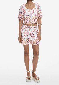 Hvid crop top med røde og lilla blomstermønstre, korte puffede ærmer og snøredetalje foran. Kombineret med matchende mønstrede shorts.