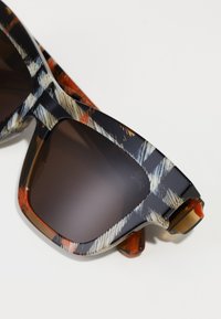 Gafas de sol con un marco de diseño en negro, marrón y crema, que presentan lentes oscuros y un logo prominente en un lado.