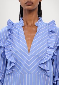 Ljusblå klänning med vertikala vita ränder, som har volangdetaljer längs kragen och ärmarna samt en djup V-ringning.