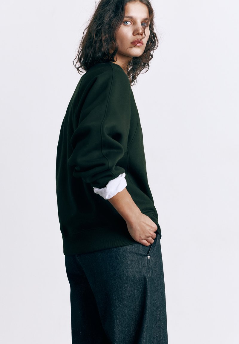 Mladić s kovrčavom kosom u crnoj majici s bijelim rukavima i tamnim trapericama, stoji bočno na bijeloj pozadini.