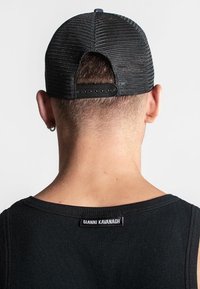 Gorra de malla negra con cierre ajustable tipo snapback, tela texturizada y una etiqueta que dice "GIANNI KAVANAGH" en la parte posterior. Se lleva con una blusa sin mangas negra.