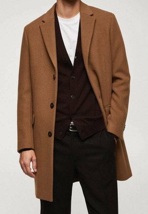 Homme portant un pardessus camel, un cardigan marron foncé boutonné, une chemise blanche et un pantalon noir, une main dans la poche et l'autre le long du corps.