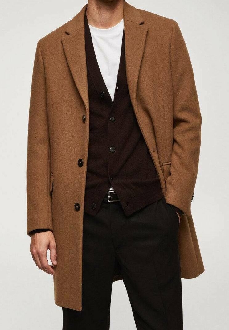 Homme portant un pardessus camel, un cardigan marron foncé boutonné, une chemise blanche et un pantalon noir, une main dans la poche et l'autre le long du corps.