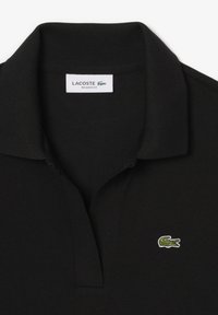 Svart polo med ribbad krage, med en liten grön Lacoste-logga broderad på vänster bröst. Tillverkad av mjukt, texturerat tyg.
