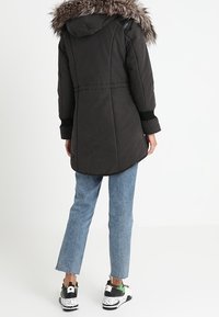 Parka nera con cappuccio a pelo, caratterizzata da una vita cinata e maniche lunghe, abbinata a jeans azzurri chiari e sneaker multicolori.