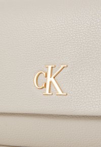 Clutch in pelle grigia chiara con texture, caratterizzata da un logo "ck" in tonalità oro. Il design è semplice, con una superficie liscia e linee pulite.