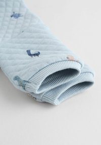 Lys blå quilted stof med ribbede manchetter, der har små dinosaur mønstre i forskellige farver. Blød tekstur og hyggeligt design.