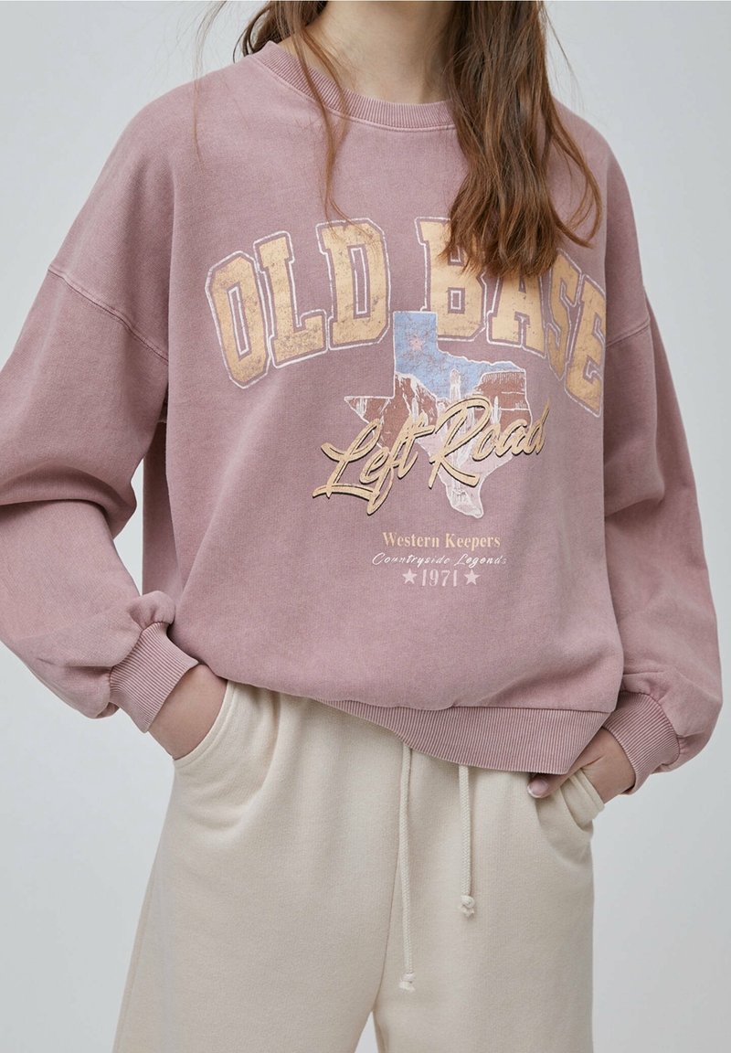 Personne portant un sweat-shirt mauve avec le texte et le graphique « OLD BASE Left Road », associé à un pantalon crème à cordon, mains dans les poches.