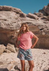 Világos rózsaszín, rövid ujjú blúz texturált díszítéssel, kék, trópusi mintás sorttal, amelynek a derékrészén zsinóros megkötő található, homokos tengerparti környezetben.