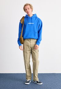 Sudadera con capucha azul y logo blanco, pantalones cargo beige con bolsillos y zapatos negros con cordones blancos, en una persona que está de pie en el interior.