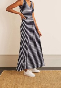 Marinblå och vitrandig, ärmlös maxiklänning med V-ringning och figursydd midja. Bärs med vita sneakers på en matta av naturfibrer.