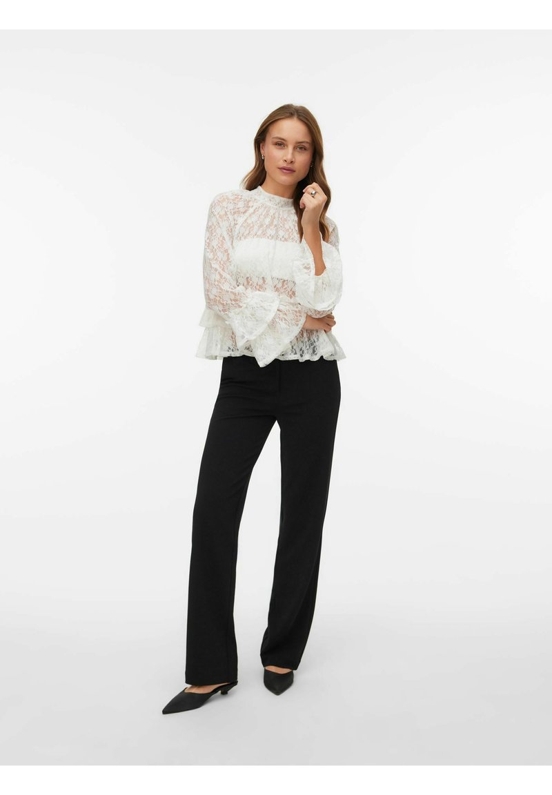 Blonderopp i hvitt med høy hals og vide ermer, kombinert med svarte bukser med rette ben og spisse svarte sko.