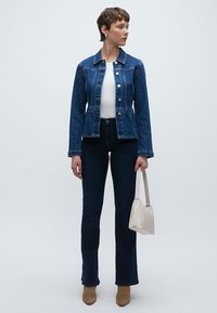 Jachetă din denim închis la culoare cu închidere cu nasturi, bluză albă și blugi închiși la culoare, cu talie înaltă. Cizme bej până la gleznă și o geantă mică, deschisă la culoare, completează ținuta.