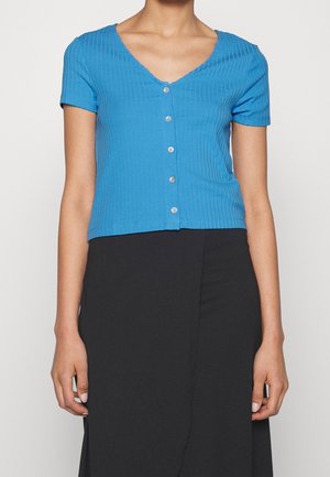 Blauw geribd shirt met korte mouwen, een V-hals en vijf knopen aan de voorkant; aansluitende pasvorm en gladde textuur. Draagt bijpassende zwarte rok.