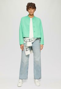 Gepolsterte, mintgrüne Jacke mit rundem Halsauschnitt und Druckknöpfen, kombiniert mit einem weißen Shirt, hellblauen Jeans und weißen Sneakers.