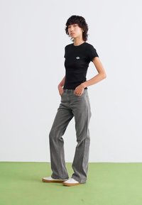 Camiseta negra ajustada con un pequeño logo, combinada con pantalones vaqueros grises holgados. El modelo lleva zapatillas blancas con suelas marrones, de pie contra un fondo liso.