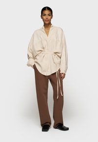 Lichtbeige, oversized shirt met een voorste knoopdetail en lange mouwen; gecombineerd met losse, chocoladebruine broek en platte zwarte schoenen.