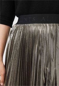 Primo piano di una gonna plissettata color oro metallizzato con cintura elastica nera e top nero a maniche corte, con il braccio sinistro e la mano in mostra.