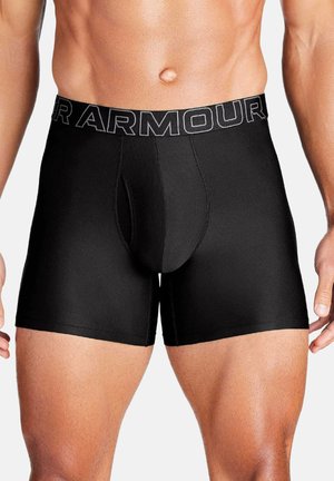 Männlicher Oberkörper, bekleidet mit schwarzen Under Armour Boxershorts mit gebrandetem Bund, zeigt muskulösen Bauch und Oberschenkel vor weißem Hintergrund.