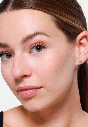 Close-up van het gezicht van een vrouw met lichte huidskleur, die kleine zilveren studoorbellen draagt. Ze heeft een natuurlijke make-up, een gladde huid en steil haar.