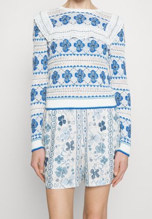 Femme portant un pull en tricot blanc et bleu avec des motifs floraux et un short assorti à motifs floraux, debout devant un fond uni.