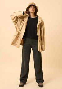 Jasno beżowa wodoodporna kurtka z regulowanym pasem, noszona na czarnym swetrze, w połączeniu z ciemnymi szerokimi spodniami i szarymi butami.
