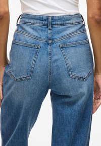 Jeans en denim bleu taille haute avec une finition délavée, dotés de deux poches arrière et d'une fermeture éclair, montrés de l'arrière.
