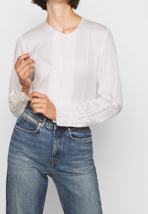 Femme portant un chemisier blanc à manches longues et un jean bleu taille haute, ajustant le poignet de sa manche gauche.