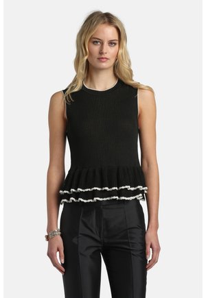 Vrouw draagt een mouwloze zwarte top met ribbelstructuur en gelaagde ruches aan de zoom met een witte rand, gecombineerd met zwarte satijnen broek en een kralenarmband.