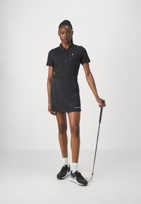Černé polo šaty s límečkem, krátkými rukávy a bočním rozparkem, v kombinaci s černými sportovními botami a golfovou holí.