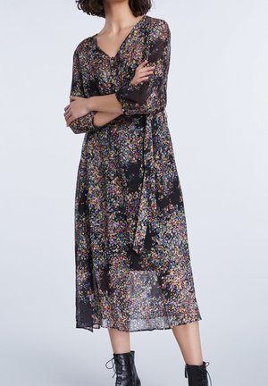 Femme portant une robe midi noire à fleurs avec des manches trois-quarts et des bottines noires à lacets, bras croisés devant un fond uni.