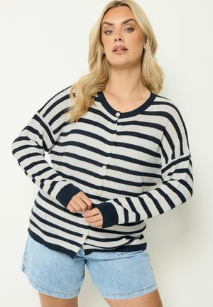 Donna bionda con cardigan a righe bianche e nere con bottoni e pantaloncini di jeans azzurri, in piedi davanti a uno sfondo neutro.