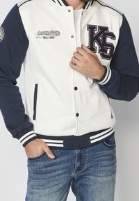 Veste varsity blanche et marine avec des poignets et un ourlet côtelés rayés. Présente un texte brodé et des détails de poche sur le côté.