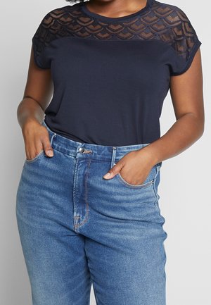 Femme portant un haut bleu marine foncé avec un motif en dentelle sur les épaules et un jean bleu taille haute, mains dans les poches, sur fond uni.