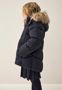 Doudoune noire avec capuche en fausse fourrure. Comprend un design matelassé, des poches avant et une fermeture éclair. Portée par-dessus une jupe sombre.