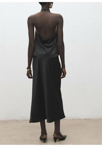 Haut cache-cou en satin noir avec dos ouvert, associé à une jupe maxi noire fluide, dotée d'une texture lisse et d'un design minimaliste.