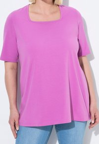 Triko s krátkým rukávem v jasně růžové barvě, vyrobené z měkké tkaniny. Má čtvercový výstřih a volný střih, kombinované se světle modrými džíny.