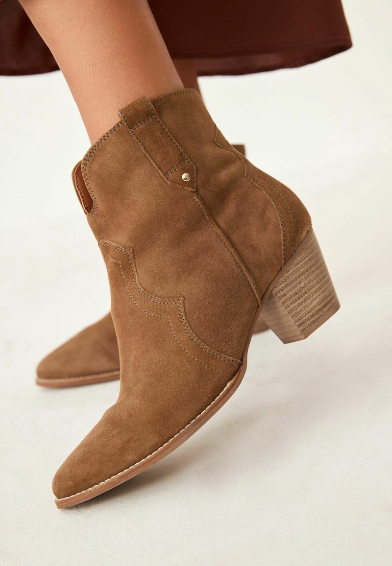Bottines en daim marron avec talon en bois empilé, couture détaillée et languette, portées avec un vêtement marron.