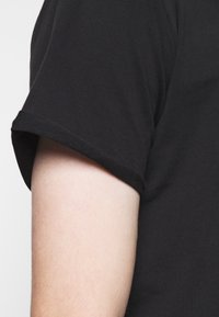 Černé tričko s krátkým rukávem, hladké na dotek a s přesně padnoucím manžetovým zakončením, ukazující část světle pleti ruky.