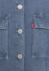 Veste en jean devant avec des boutons en métal et deux poches à rabat, avec une étiquette rouge de la marque Levi's sur la couture de la poche droite.