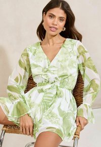 Ziedu kleita ar zaļu un baltu botānisku rakstu, v-veida dekoltē, ar ruffled detaļām, garām caurspīdīgām piedurknēm un saitēm viduklī.