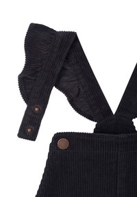 Black corduroy overalls feature flared, ruffled straps with bronze snap closures and a decorative logo patch on the pocket.