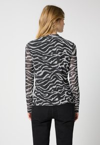 Langärmliges, transparentes Oberteil mit einem schwarz-grauen Zebra-Druck. Die Rückansicht zeigt die figurbetonte Passform und die leichte Textur. Getragen mit schwarzen Hosen.